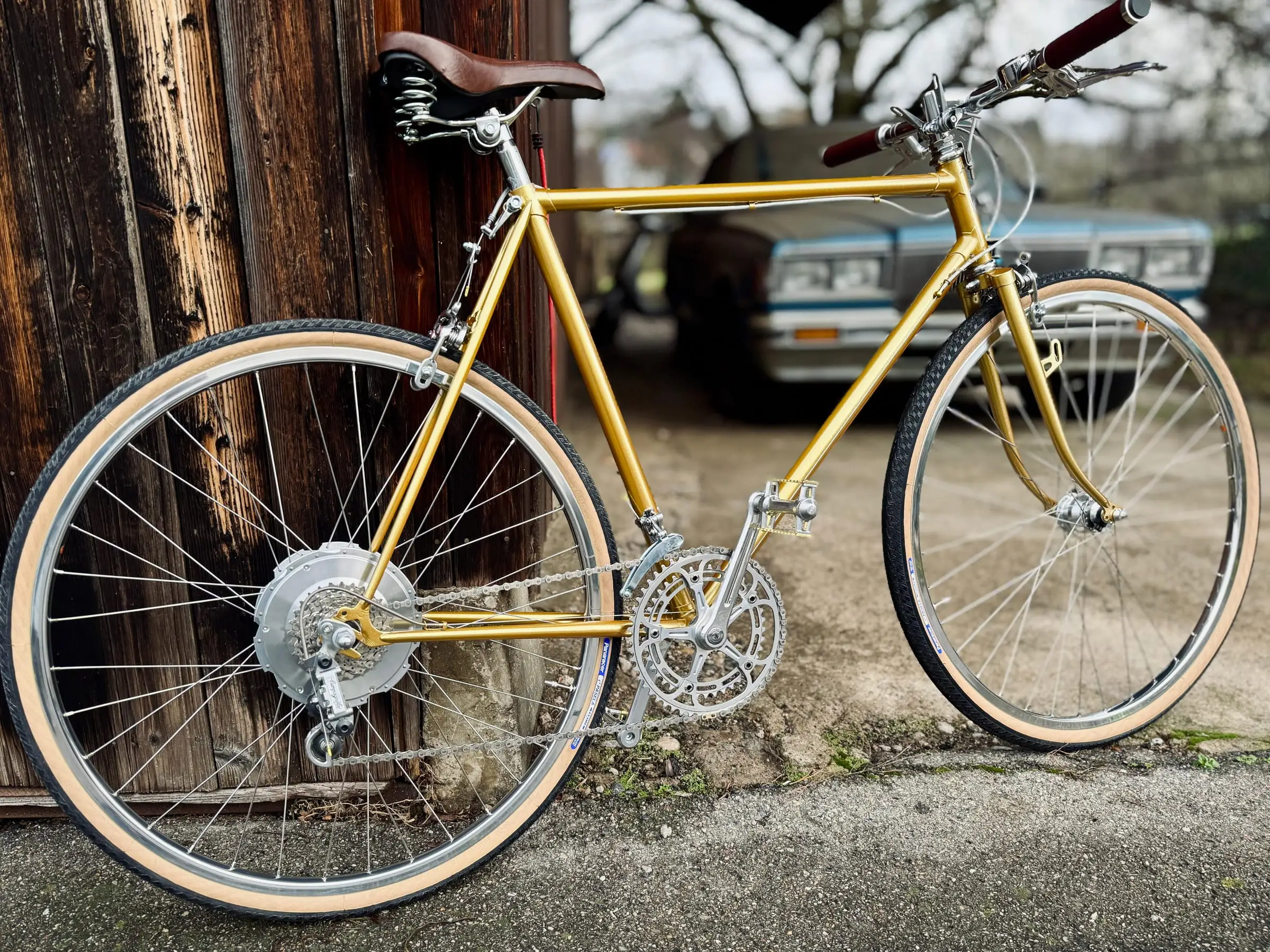 Goldenes Cenci-Velo mit Zehus-Nabenmotor im Hinterrad, Ledersattel und cremefarbenen Reifen, vor der Holzwand der Werkstatt
