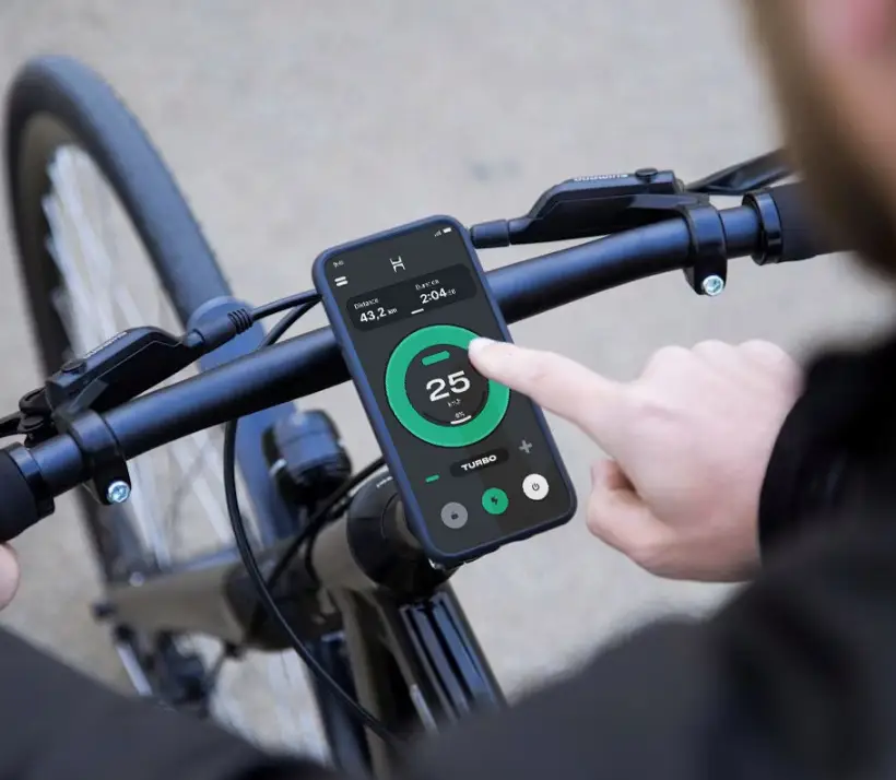 Ein Finger tippt auf das am Velolenker montierte Smartphone: Die Zehus App zeigt 25 km/h im Turbo-Modus, dahinter Vorderrad und Bremshebel